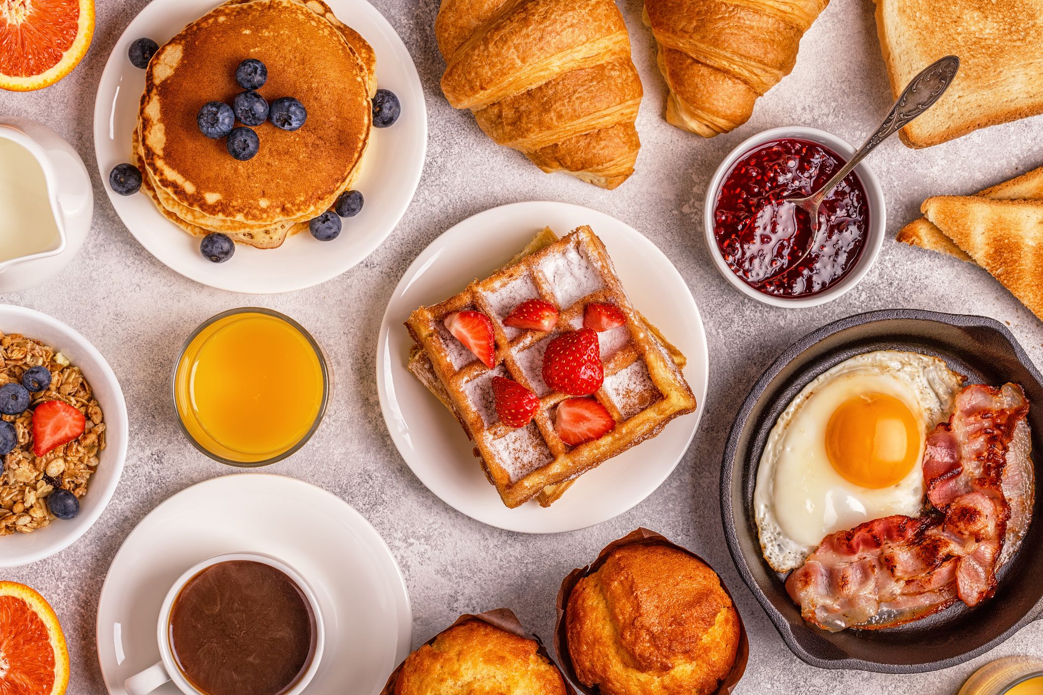An arrangement of waffles, fruit, croissants, eggs, bacon, pancakes, and other breakfast foods spread on a table.